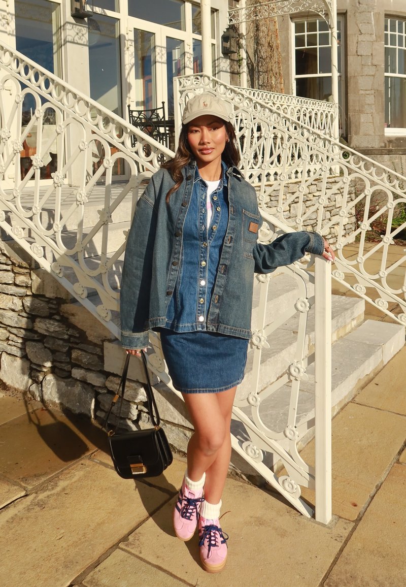 Conjunto de jaqueta e saia de denim em azul, com uma mala preta e ténis cor-de-rosa. O boné é bege com um detalhe de logótipo. O fundo brilhante apresenta escadas.