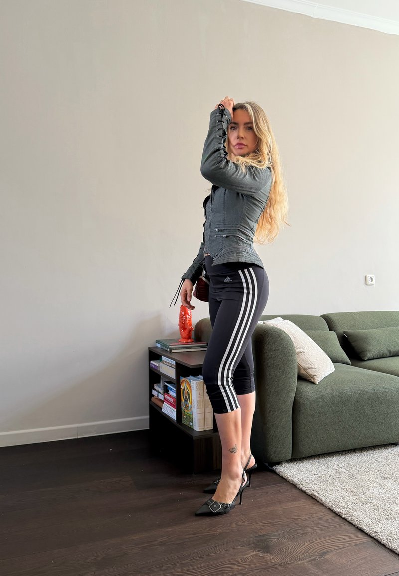 Femme aux longs cheveux blonds portant une veste grise, des leggings Adidas noirs et des talons noirs, debout à l’intérieur près d’un canapé vert et d’une bibliothèque.