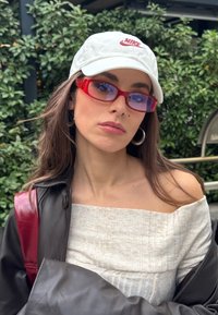 Jeune femme portant une casquette blanche Nike, des lunettes de soleil rouges translucides, des boucles d'oreilles créoles, un haut blanc à épaules dénudées et une veste en cuir noire à l'extérieur.