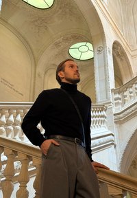 Osoba stoi obok ozdobnej klatki schodowej. Ma na sobie czarny golf i szare dopasowane spodnie. Jasnozielone świetliki oświetlają przestrzeń.