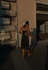 Fekete ujjatlan felső, hátsó megkötővel, sötét farmer rövidnadrág és papucs. Mintás kézitáska a kezében, egy textúrált kőfal mellett sétál.