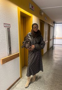 Hnědá kožená bunda s kožešinovým límcem, dlouhá vzorovaná sukně a béžové boty. Stojí v hale u žlutých dveří výtahu.