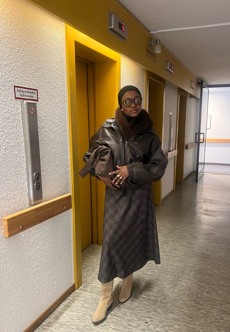 Braune Lederjacke mit Pelzkragen, langer gemusterter Rock und beigefarbene Stiefel. Steht in einem Flur mit gelben Aufzugtüren.
