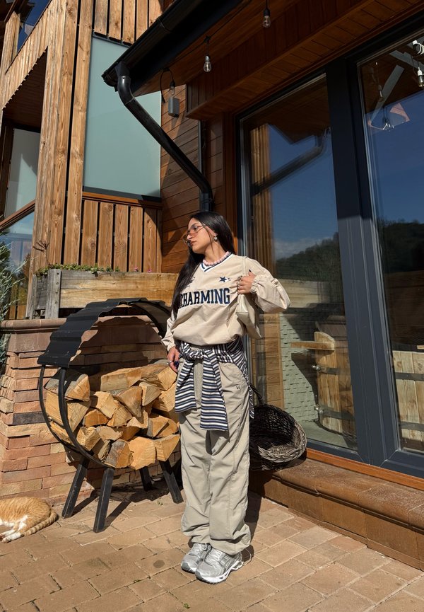 Joven mujer con sudadera beige "Charming" y pantalones cargo, de pie junto al exterior de una casa de madera con leña apilada y un gato naranja durmiendo cerca.