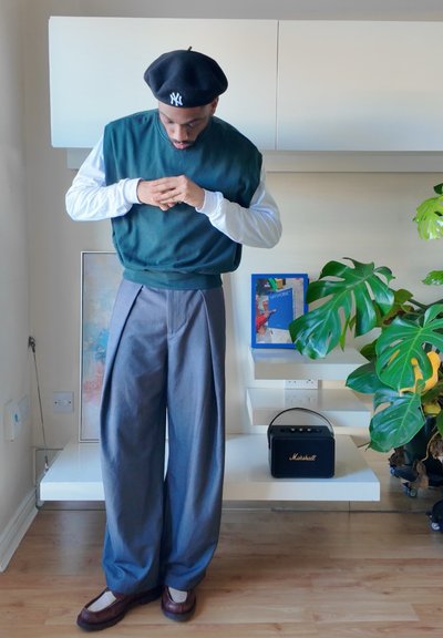 Sudadera de forro polar verde sobre una camisa blanca de manga larga, combinada con pantalones grises plisados y zapatos marrones. De fondo hay una planta y decoración.