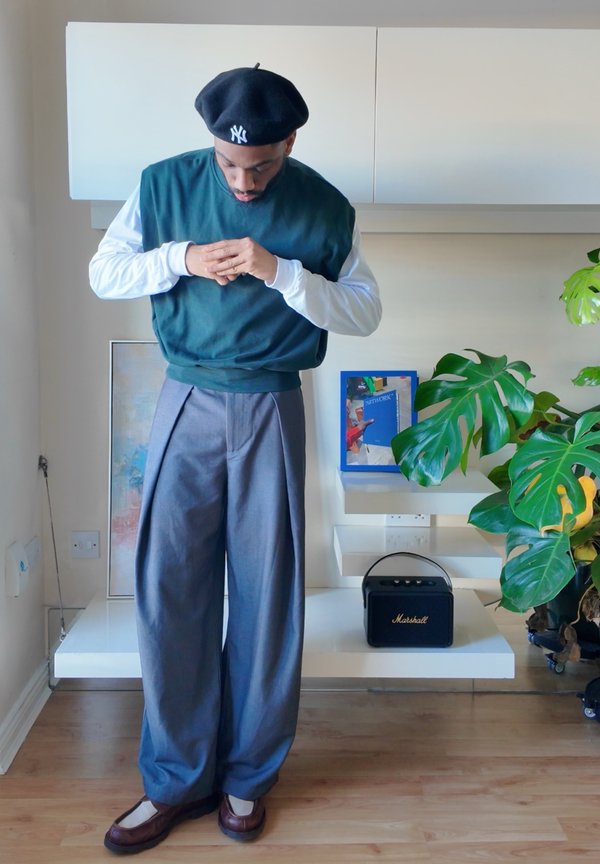 Roheline fliisist dressipluus, mida kantakse valge pika varrukaga särgi peal, kombineeritud hallide volditud pükstega ja pruunide kingadega. Taustal on taim ja dekoratsioonid.