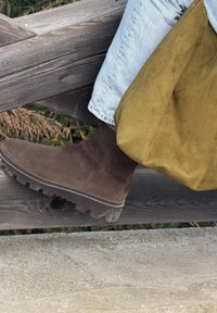 Botas de tornozelo em camurça castanha com uma sola espessa e texturada, apoiadas numa cerca de madeira. Uma bolsa em camurça verde está pendurada nas proximidades, parcialmente visível.