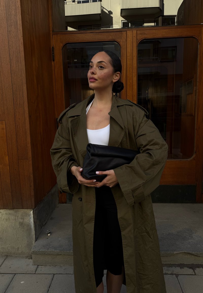 Femme aux cheveux foncés en chignon portant un trench-coat olive oversize et un haut blanc, tenant une grande pochette noire, debout devant des portes en bois.