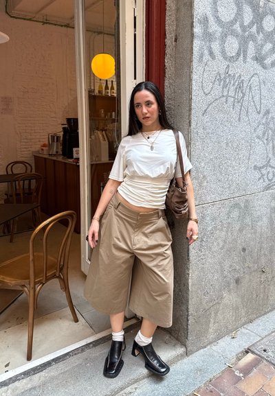 Camiseta blanca fruncida, pantalones anchos beige, bolso marrón, botines negros de tacón con recortes y calcetines de diseño. Entorno urbano.