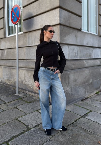Camisa negra corta con cremallera, jeans de pierna ancha azul claro, cinturón negro con hebilla plateada y zapatos negros, complementados con gafas de sol y un bolso.