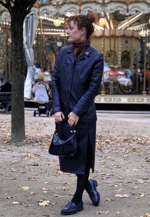 Chaqueta de cuero negra y falda con una abertura lateral. Combinada con una bufanda de estampado y un bolso negro brillante. Zapatos oscuros completan el look.