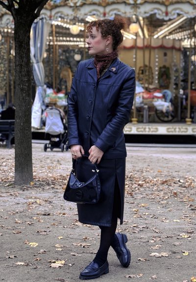 Chaqueta de cuero negra y falda con una abertura lateral. Combinada con una bufanda de estampado y un bolso negro brillante. Zapatos oscuros completan el look.