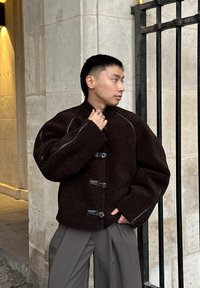 Brun fleecejakke med høy krage, svart kant og trykknapper. Stylet med løse grå bukser, mot en bakgrunn av steinvegg.