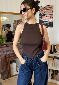 Jeune femme portant des lunettes de soleil, un haut marron sans manches et un pantalon bleu taille haute, se tenant les mains dans les poches dans une pièce avec des livres et une affiche.