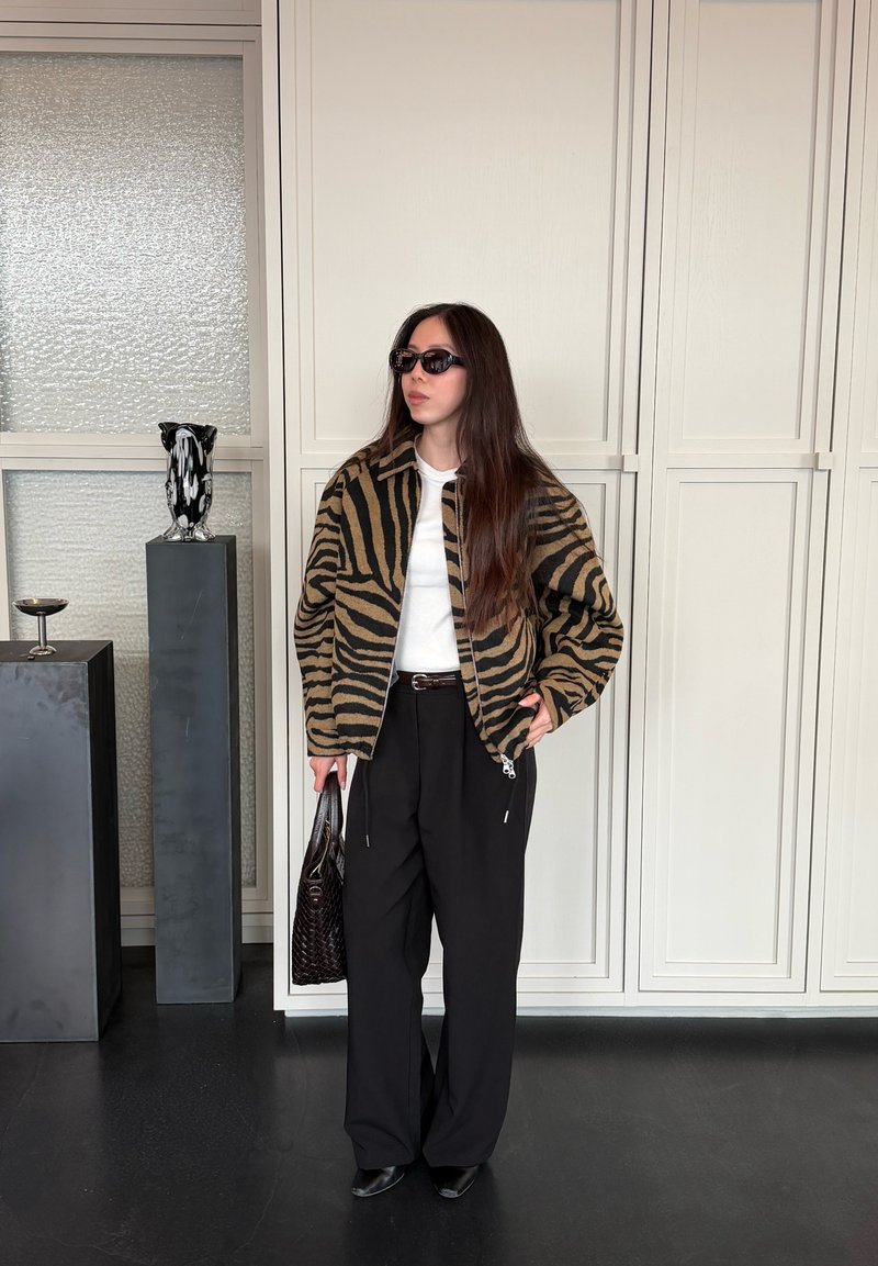 Femme portant une veste à imprimé tigre, chemise blanche, pantalon noir, lunettes de soleil, tenant un sac à main noir texturé à l’intérieur près de vases décoratifs.