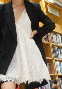 Juodas švarkas ant balto suknelės su nėriniu, su V formos iškirpte, sluoksniuota medžiaga ir laisvu siluetu. Fone matomos knygų lentynos.