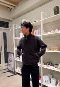 Homme aux cheveux bouclés foncés portant une veste zippée debout dans l'intérieur d'un magasin, avec des étagères présentant divers produits emballés derrière lui.