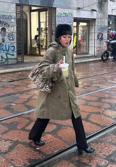 Abrigo impermeable verde, pantalones negros, bolso con estampado, sosteniendo té de burbujas. Calle empedrada mojada, fondo de tienda urbana con graffiti.