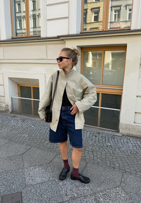 Woman wearing black sunglasses, beige jacket, black top, denim shorts, and black shoes standing on a cobblestone street in front of a building.