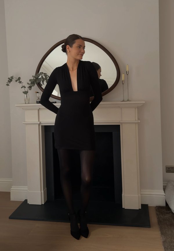 Femme en robe noire et collants, debout les mains derrière le dos devant une cheminée blanche avec un miroir ovale et des objets décoratifs.