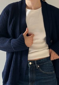 Temno moder riban cardigan z ovratnikom in zapiranjem s sprednjimi gumbi, oblečen preko belega ribanega srajca in v kombinaciji s temno modrimi kavbojkami.