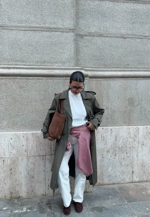 Persona con abrigo largo color oliva, camisa blanca, pantalones blancos, zapatos marrones, sosteniendo bolso marrón, con camisa roja a cuadros atada alrededor de la cintura, de pie al aire libre.