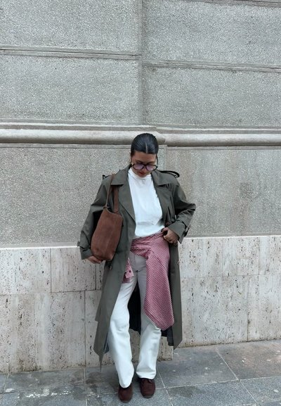 Persona con abrigo largo color oliva, camisa blanca, pantalones blancos, zapatos marrones, sosteniendo bolso marrón, con camisa roja a cuadros atada alrededor de la cintura, de pie al aire libre.
