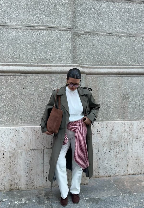 Person trägt langen olivgrünen Trenchcoat, weißes Hemd, weiße Hose, braune Schuhe, hält eine braune Tragetasche, mit einem rotkarierten Hemd um die Taille gebunden, steht im Freien.