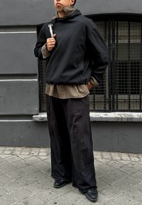 Czarna, oversize bluza z wysokim kołnierzem, nałożona na brązową koszulę w kratę, w połączeniu z luźnymi czarnymi spodniami i czarnymi trampkami.