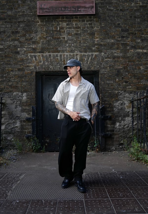 Joven con tatuajes, vestido con gorra, camisa de cuadros, camiseta blanca y pantalones negros, de pie frente a una vieja pared de ladrillos con una puerta oscura.