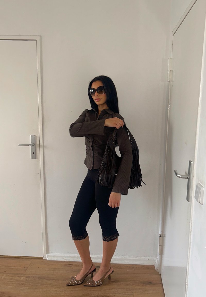 Femme portant des lunettes de soleil foncées, une veste marron, des leggings noirs courts avec bordure en dentelle, des talons marron, et tenant un sac à main noir à franges à l'intérieur.