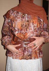 Cinturón marrón ajustando una blusa floral multicolor con volantes en tonos morado, naranja y beige, usada sobre una falda blanca de encaje. Anillos decorativos.
