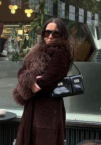 Femme portant un manteau marron foncé bordé de fourrure et des lunettes de soleil, tenant un sac à main noir, debout devant un café avec une fenêtre en verre réfléchissant en arrière-plan.