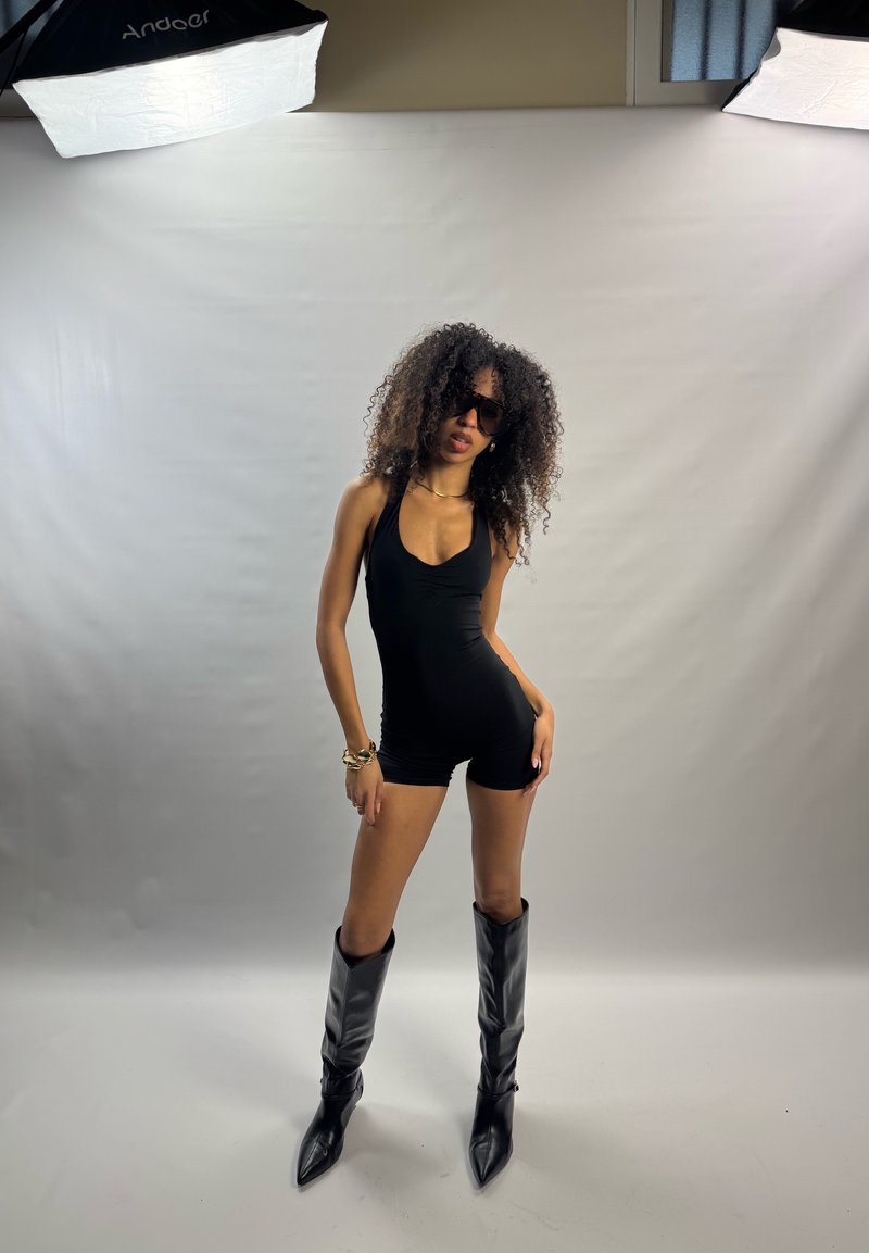 Femme aux cheveux bouclés portant une combinaison noire sans manches, des bottes pointues noires, des lunettes de soleil et des bijoux en or, posant dans un studio avec un fond blanc et des lumières.