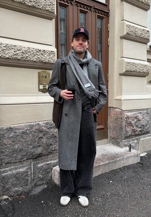Hombre con abrigo gris y bufanda, gorra negra y zapatillas, sosteniendo una taza de café, de pie frente a la entrada de un edificio de piedra.