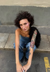 Ženska s kodrastimi lasmi, tetovažami na desni roki, oblečena v brezramen jeans kombinezon in črne čevlje, sedi na robniku ob sivi steni.