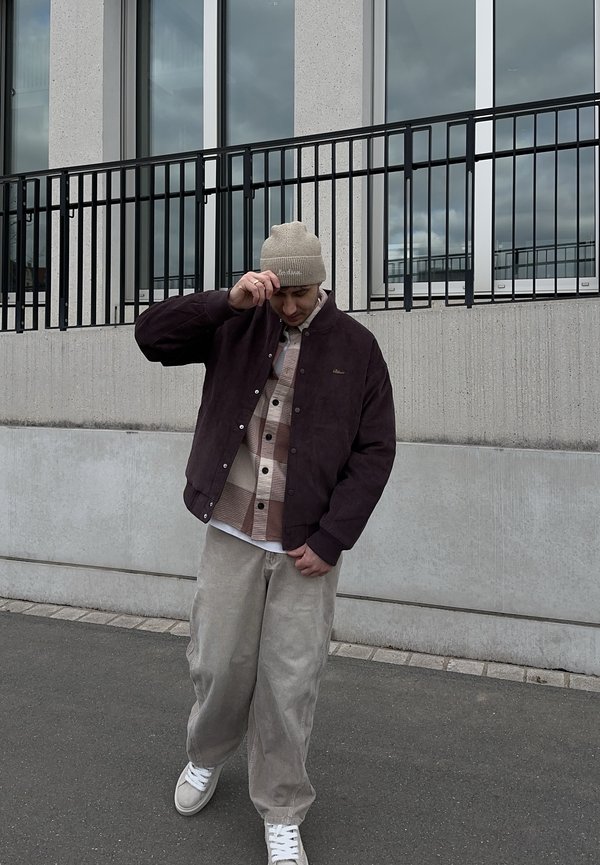 Homme portant un bonnet beige, une veste sombre, une chemise à carreaux et un pantalon large beige ajuste son chapeau tout en se tenant sur le trottoir près d'un mur en béton avec des fenêtres.
