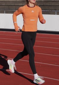 Tenue de course comprenant un haut à manches longues orange vif avec des accents blancs et des leggings noirs. Les chaussures sont blanches avec des accents verts et roses.