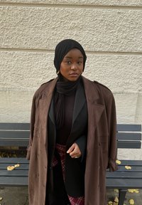 Cappotto doppiopetto in suede marrone sovrapposto a un top nero e a una sciarpa. Orecchini a cerchio dorati. Lo sfondo presenta un muro texturizzato e una panchina.