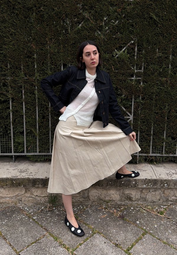 Femme aux cheveux foncés porte une veste noire, un pull blanc, une jupe beige et des chaussures noires à pois blancs, posant près d'une haie et d'une clôture métallique.