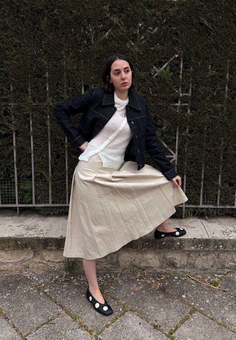 Femme aux cheveux foncés porte une veste noire, un pull blanc, une jupe beige et des chaussures noires à pois blancs, posant près d'une haie et d'une clôture métallique.