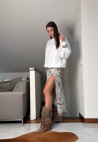 Vit överdimensionerad sweatshirt, blommig genomskinlig kjol med hög slits, och bruna fransade stövlar. Släta texturer och flödande tygdetaljer.
