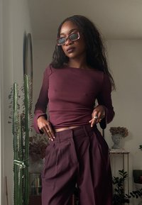 Mørkerød ribbet langærmet top med udslåede manchetter kombineret med højtaljede, plisserede mørkerøde bukser. Tilbehør inkluderer lyserøde solbriller.