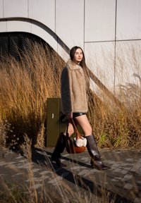 Femme marchant en plein air, portant un manteau en fausse fourrure beige, un short en cuir noir, de hautes bottes marron et tenant un sac à main marron avec des cache-oreilles blancs.