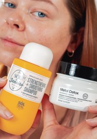 Femme tenant près de son visage une bouteille jaune de shampooing Fortifiant + Lissant et un pot blanc de masque Détox Métal.