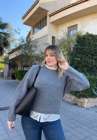 Grå strikkegenser med ballongerm kombinert med en hvit skjorte med lag-på-lag og volanger, mørke jeans og en svart skulderveske på fortauet.