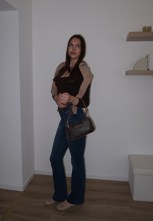 Jeune femme aux longs cheveux foncés portant un haut marron, un jean bleu, un pull beige drapé sur les épaules, des ballerines beiges, tenant un sac à main marron à l'intérieur.