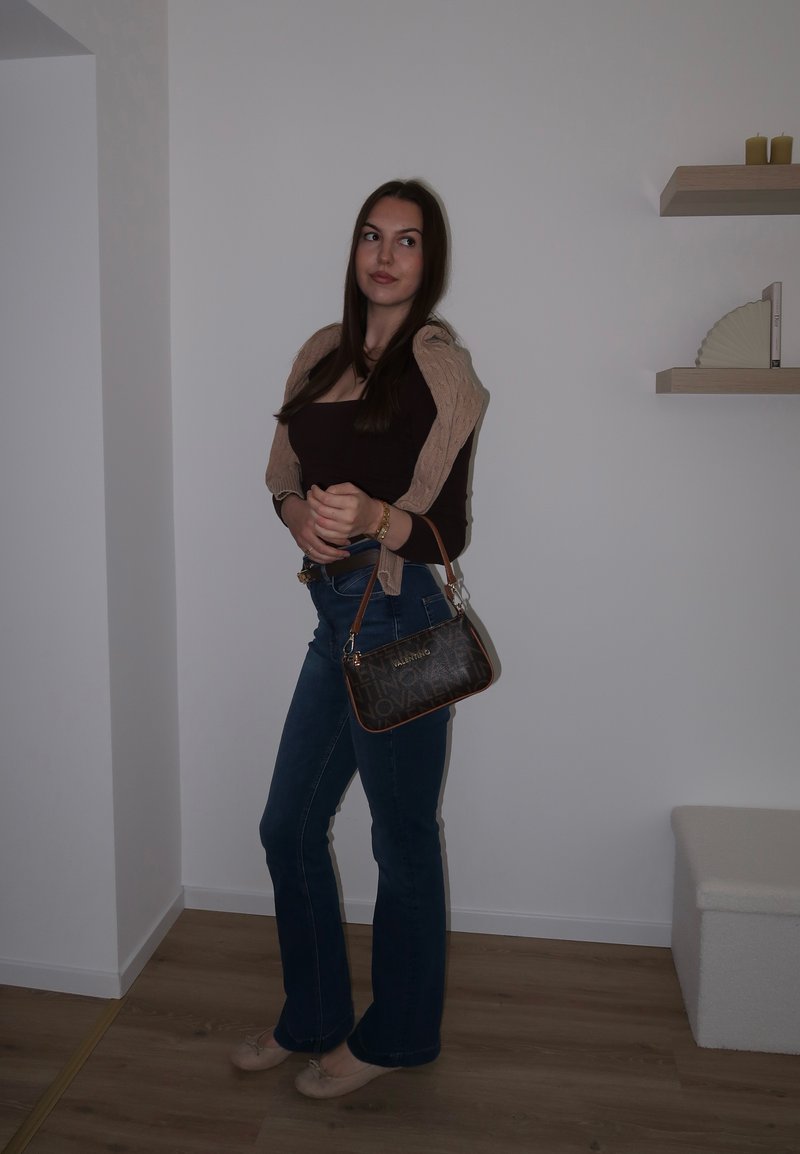 Jeune femme aux longs cheveux foncés portant un haut marron, un jean bleu, un pull beige drapé sur les épaules, des ballerines beiges, tenant un sac à main marron à l'intérieur.