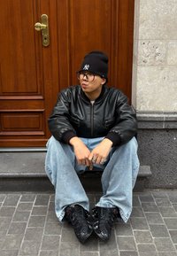 Jeune personne portant une veste en cuir noire, un bonnet noir et un jean large, assise sur des marches devant une porte en bois, regardant sur le côté avec les mains jointes.