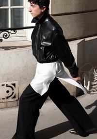 Giacca bomber in pelle nera con colletto alto, abbinata a una camicetta bianca e pantaloni neri a gamba larga. Porta una borsa beige con frange.